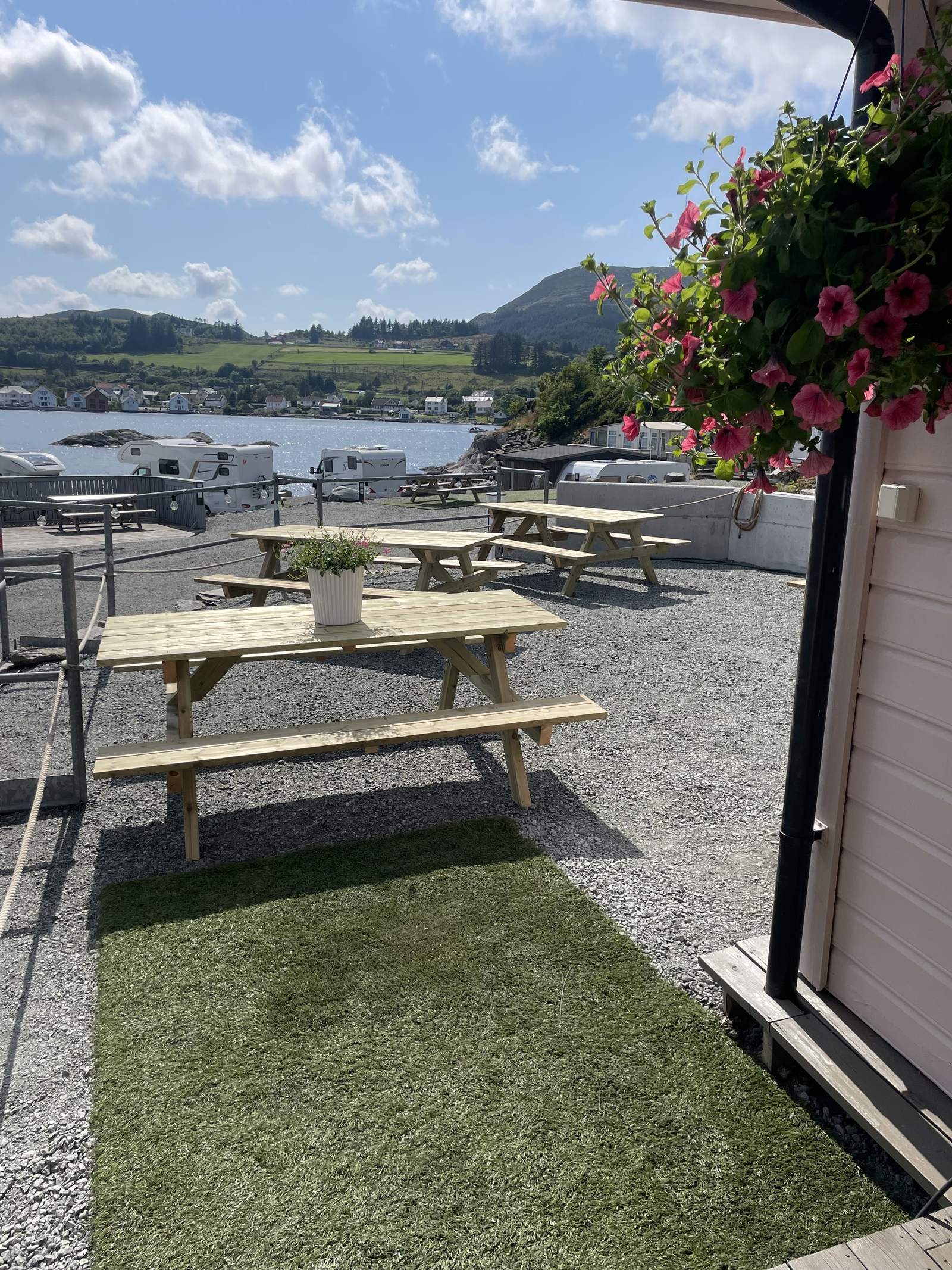 Bordbenker med blomster på plassen og utsikt mot bobiler, sjøen og grønne åser