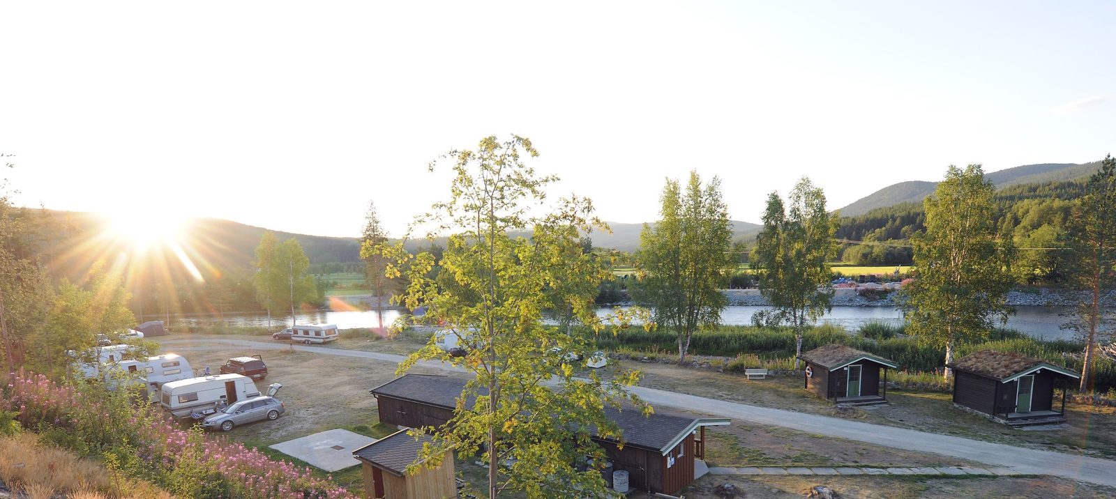 Bobiler og campingvogner på grasplass ved elva Folla, med Tronfjellet i bakgrunnen