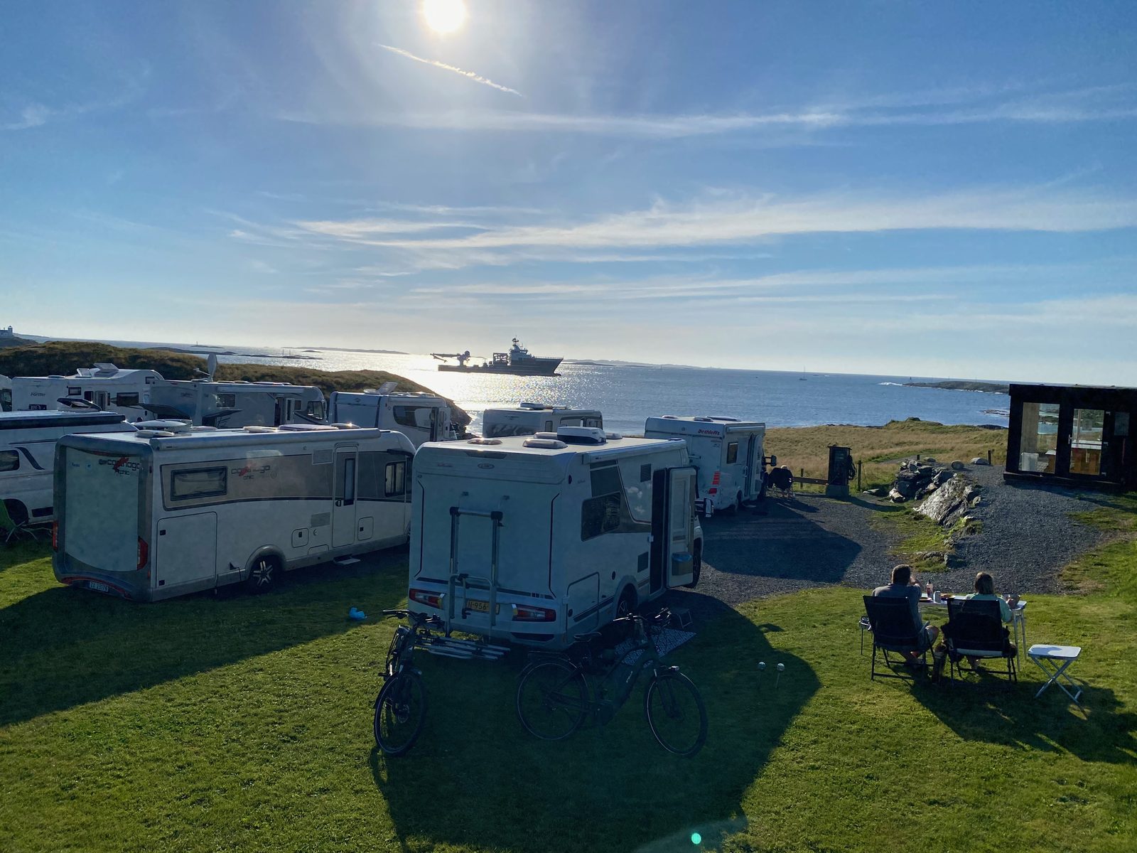 Campingvogner og bobiler nær sjøen med skip og glasshytte til høyre i kveldssol