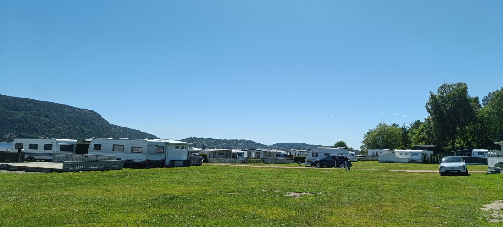Stor gresslette på Homannsberget Camping med campingvogner og biler langs kanten og skogkledde åser bak
