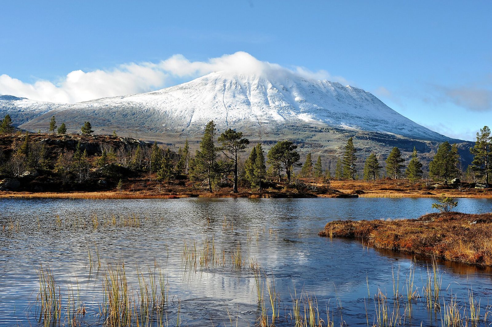 Gaustatoppen speiler seg i stille fjellvann med snø på toppen og høstfarger i forgrunnen