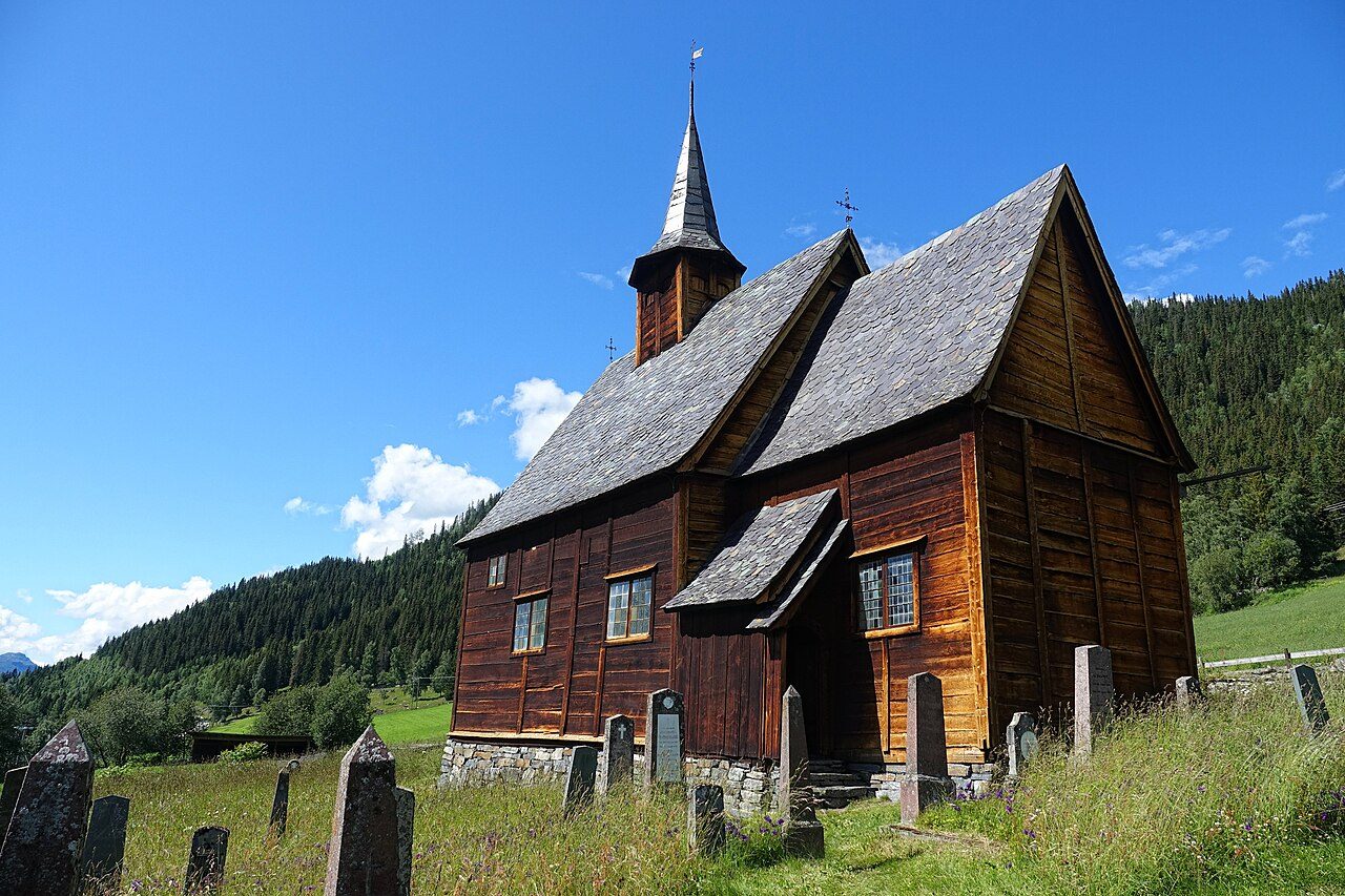Lomen stavkirke i Valdres med mørk tjærebredd tømmer og utskårne portaler mot grønt dalføre